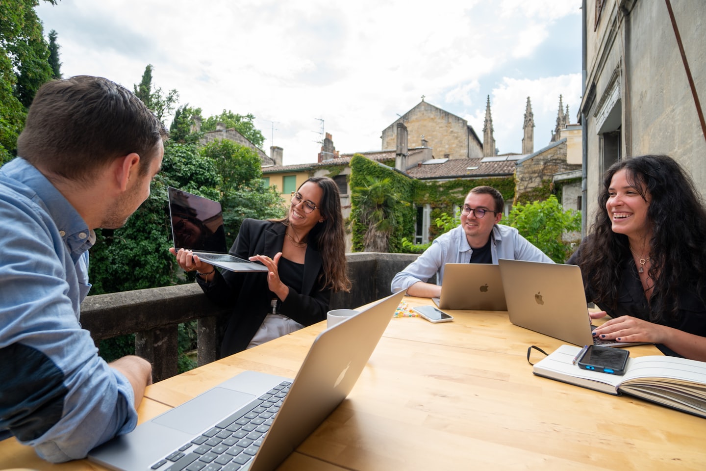 Four young professionals collaborate at an outdoor table with laptops.