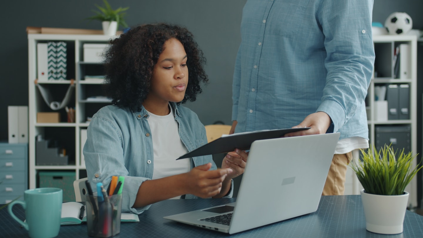Two colleagues collaborate at a desk with a laptop and folder.